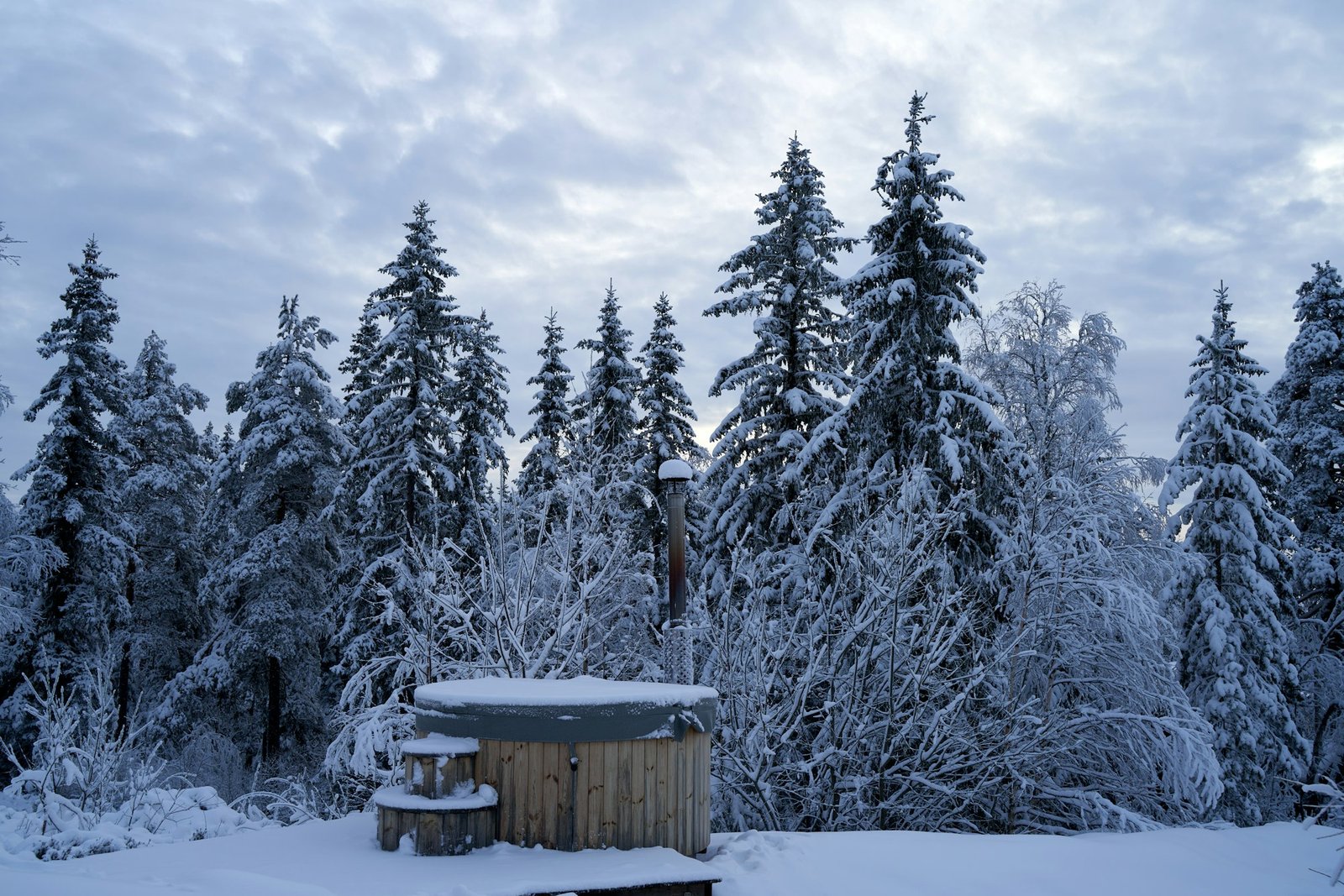 A wooden hot tub surrounded by snow-covered pine trees