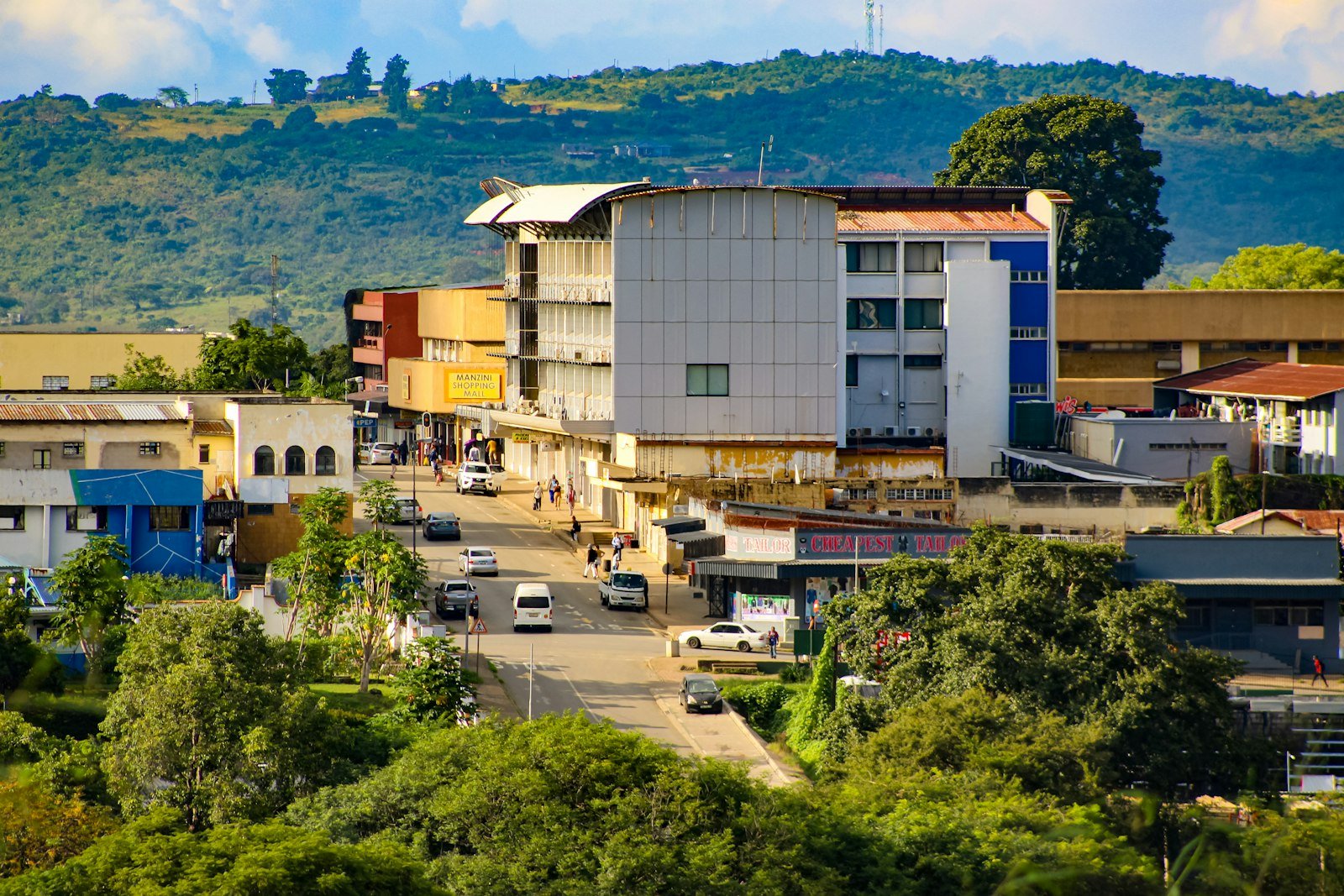 Buildings line a street with cars and green hills